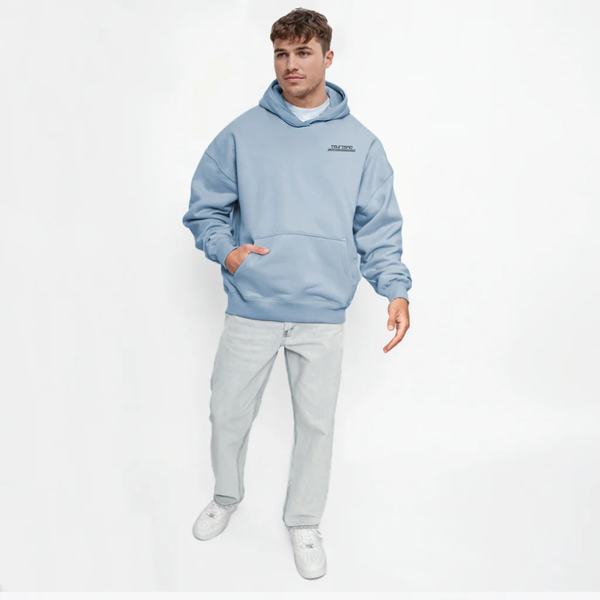 Homme portant un hoodie bleu clair TOURISMO avec jean clair, fond blanc