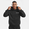 Sweat à capuche noir sans cordon pour homme, modèle "Pression Maximale" signé Tourismo Clothing. Hoodie coupe droite avec poche kangourou, capuche structurée tenue fermement par les deux mains du mannequin. Logo brodé blanc "TOURISMO" avec damier graphique sur la poitrine gauche. Fond studio blanc, cadrage buste, regard frontal intense. Vêtement streetwear inspiré de l’univers automobile, entre technicité textile et attitude déterminée. Idéal pour un look racing discret et puissant.