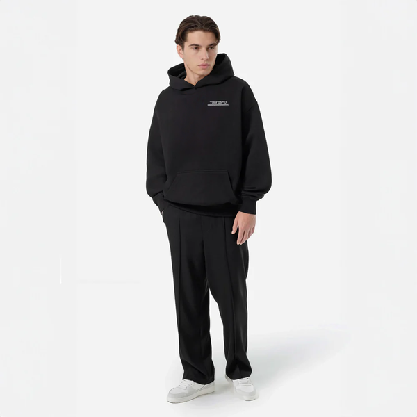 Homme portant un hoodie noir TOURISMO, regard gauche, fond blanc, pantalon habillé